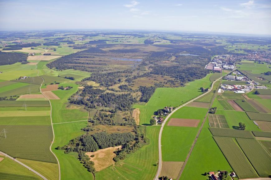 Zwischen Wiesen und Ackerflächen liegt hier ein ausgedehntes Naturschutzgebiet mit Wald, Seen und Moorflächen. Rechts oben sind die Ausläufer einer Siedlung erkennbar. Blick aus großer Höhe.