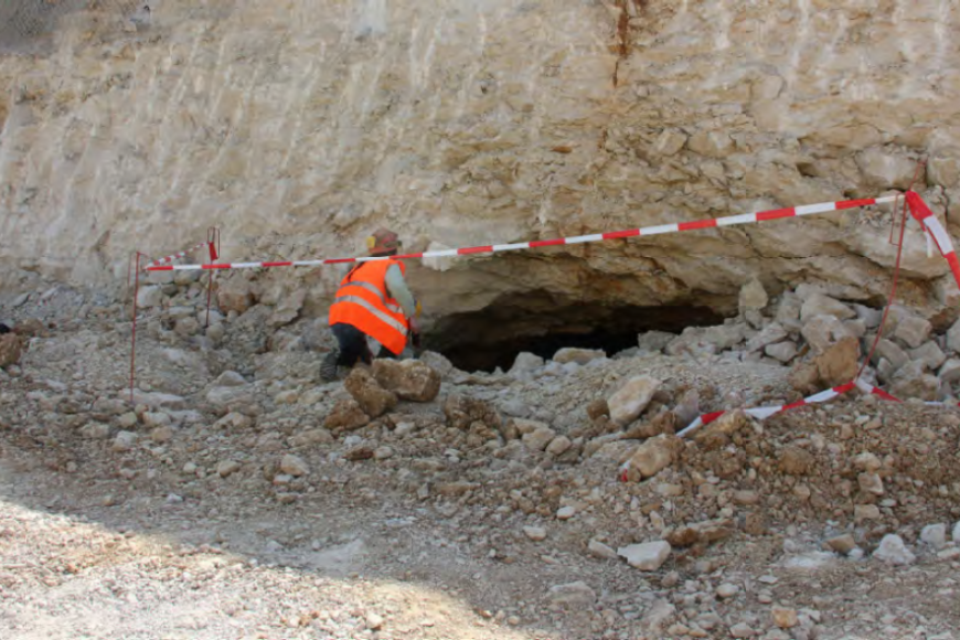 Zu sehen ist eine hohe hellbraune bis weiße Steinwand, an deren unteren Ende eine Person mit Warnweste vor einer Höhlenöffnung kniet. Um die Höhle ist Absperrband aufgehängt.