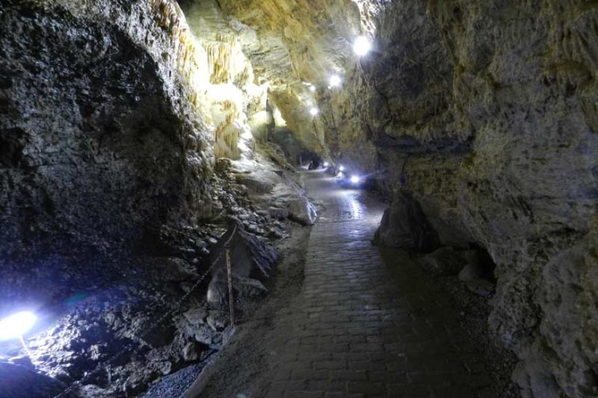 Blick in eine Tropfsteinhöhle. Zwischen links und rechts aufsteigenden Gesteinswänden führt ein schmaler, gepflasterter Weg entlang. Der hintere Teil der Höhle ist mit Lampen ausgeleuchtet.