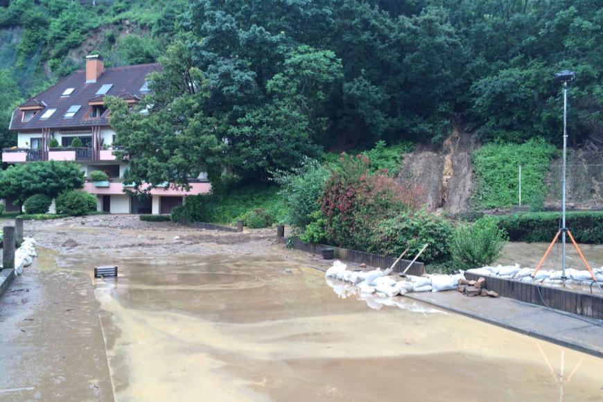 Das Bild zeigt eine von Schlamm und Wasser überspülte Straße. Die Straße führt links an einem rechts aufsteigenden steilen Waldhang entlang. Rechts sind Schlammspuren am Hang sowie Sandsäcke zu sehen.