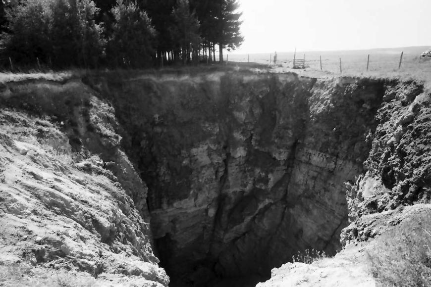 Altes Schwarzweißfoto, das den tiefen Krater einer Doline zeigt. Das freiliegende Boden- und Gesteinsmaterial befindet sich auf einer Wiese am Rande eines Waldes.