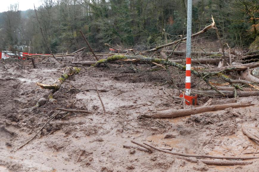 Auf dem Bild sind große Mengen an braunem Schlamm zu erkennen, aus denen mitgerissene Baumstämme und Äste ragen. Inmitten der Schlammmassen stehen zwei Laternenpfeiler, die zum Parkplatz des großen roten Gebäudes im Hintergrund gehören.