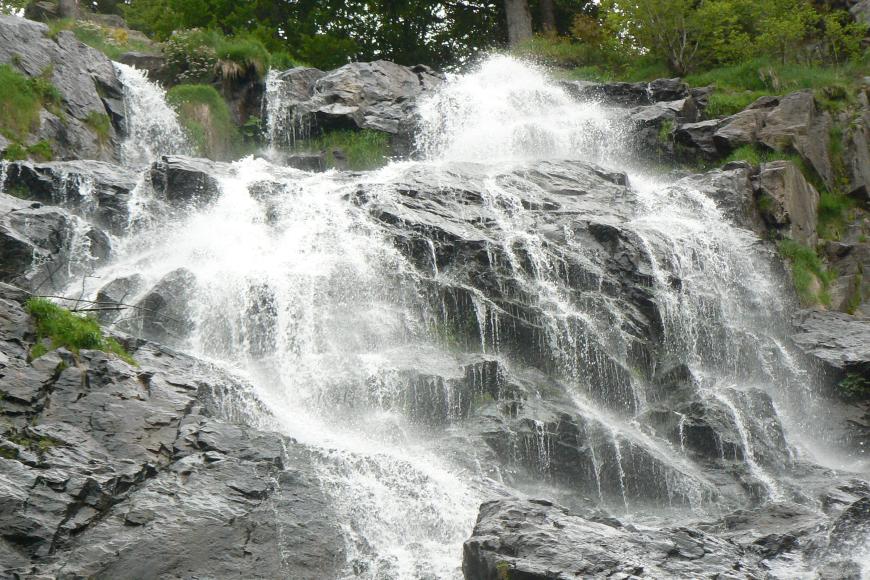Das Bild zeigt einen breiten Wasserfall, der mehrere kantige, graue Felsstufen mit weißer Gischt überspült. Am oberen Bildrand sind Bäume und Sträucher erkennbar.
