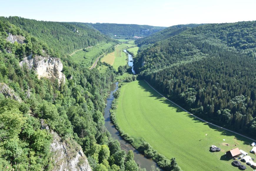 Blick aus großer Höhe über ein langgezogenes Tal mit Flusslauf. Das Tal ist hufeisenförmig von bewaldeten Bergen eingerahmt. Am höchsten Berghang links im Vordergrund stehen bleiche Felsen heraus.