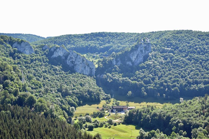 Blick auf bewaldete Berge, die sich links und rechts am Fuß eines kleinen Tals erheben. Ebenfalls links und rechts treten steile graue Felsen aus den Bergrücken hervor und bilden ein flaches V. Der Felsen rechts trägt oben mehrere Gebäude.