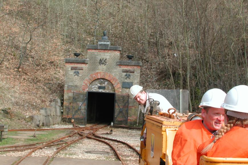 Besucherbergwerk Tiefer Stollen in Aalen-Wasseralfingen Blick auf den geöffneten, an einem Waldhang gelegenen Eingang eines Bergwerks. Im Vordergrund liegen Schienen der Grubenbahn.