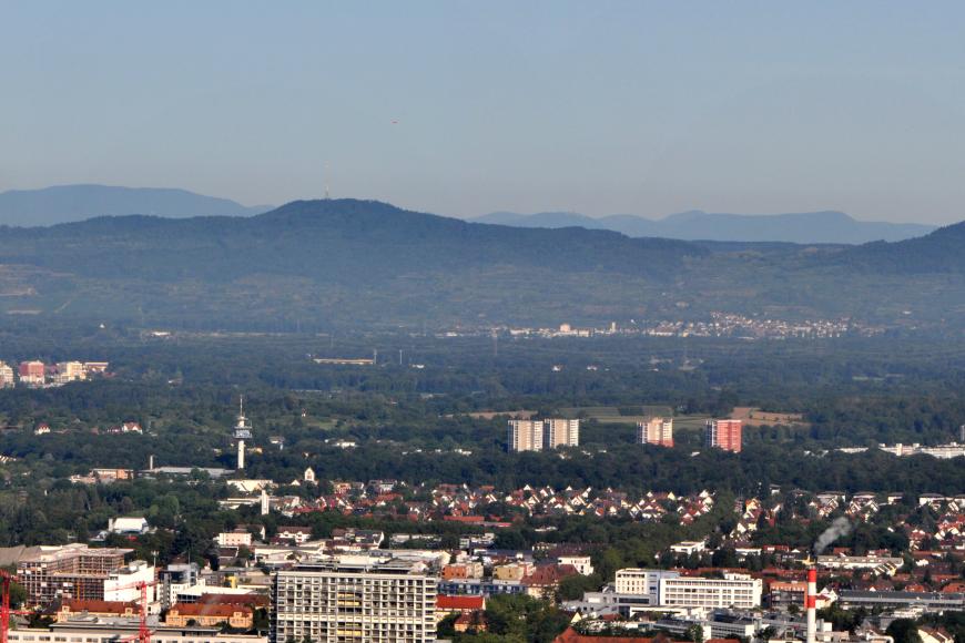 Panoramabild einer größeren Stadt mit Altstadt- und Hochhäusern. Rechts unten ist eine größere Baustelle zu erkennen. Den Hintergrund bilden bewaldete Hügel- und Bergketten.