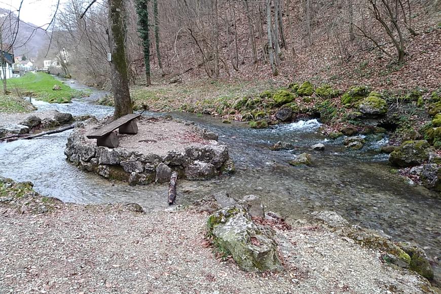 Echazquelle Von einer Ortschaft links hinten strömt ein Wasserlauf nach rechts am Betrachter vorbei. Links der Bildmitte umfließt er dabei eine kleine, von Felsgestein umrandete Insel mit Baum und Sitzbank. Das vordere Ufer ist flach, das hintere bildet ein Waldhang.