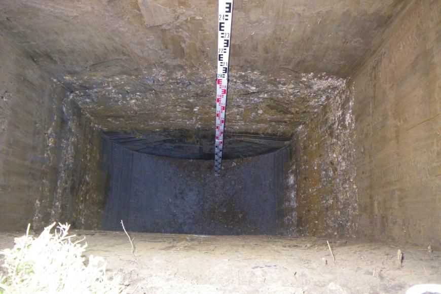 Das Foto zeigt einen rechteckigen Baggerschurf in Aufsicht. Allseitig sind ockerfarbene braune senkrechte Wände zu sehen. Im untersten Teil zeichnen sich grau kiesige und an der Basis dunkelbraune tonige Bereiche ab. An einer Wand lehnt eine Messlatte.