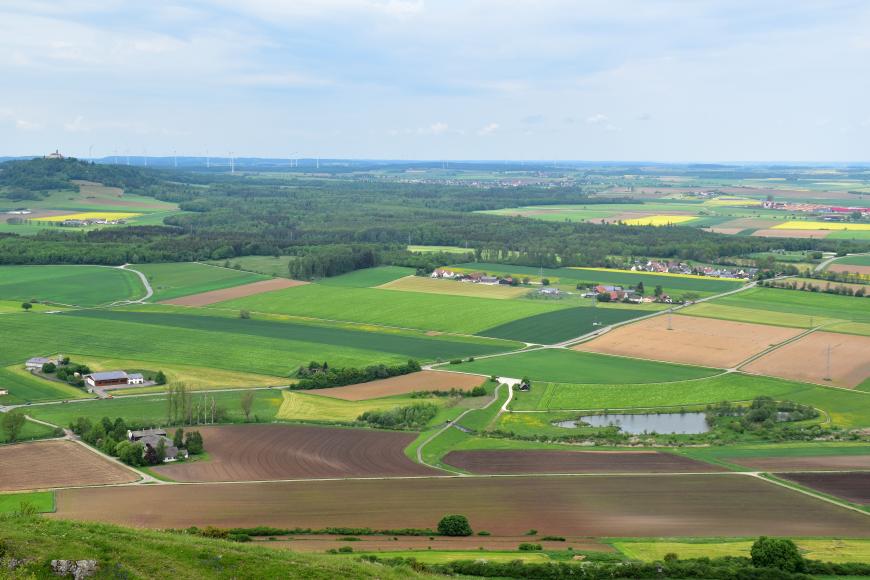 Blick aus größerer Höhe über eine flache, im Hintergrund links auch hügelige Landschaft mit größeren Acker-, Wiesen- und Waldflächen. Im Vordergrund rechts ist ein kleiner länglicher See zu erkennen. Im Hintergrund stehen Windräder.