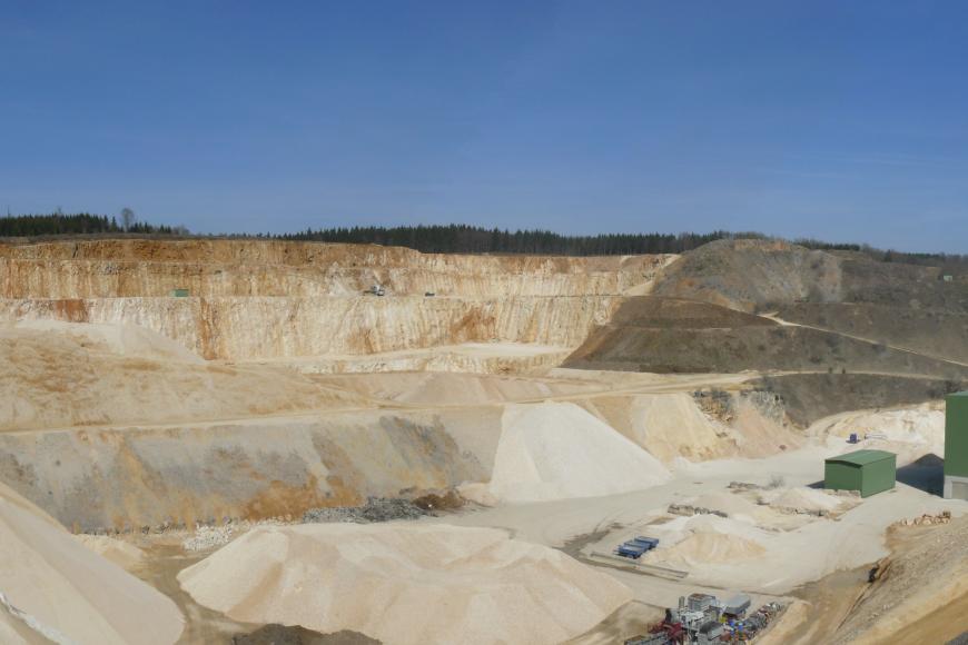 Blick in einen großen Steinbruch mit aufgehaldeten hellbeigen Splitten und Schottern auf der untersten Sohle.