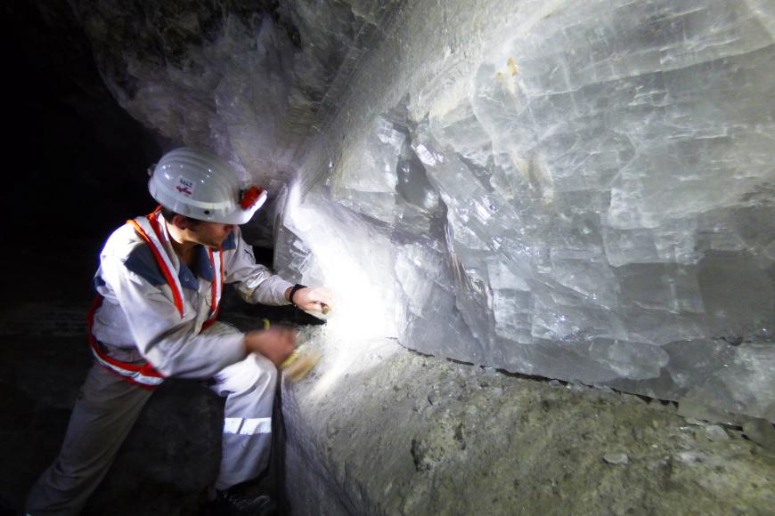 Ein Bergbauarbeiter links im Bild beschäftigt sich mit einer Wand aus großen, weißlichen Kristallen, die auf grünlichem Gestein aufliegt.