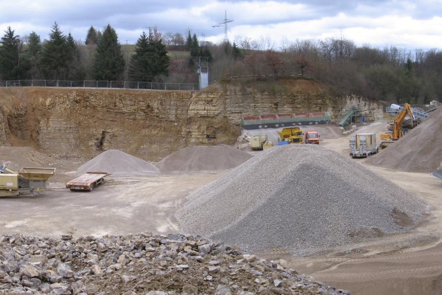 Blick von erhöhtem Standpunkt in einen großen Steinbruch. Es sind Baustellen-Fahrzeuge erkennbar. Das anstehende Gestein ist hellbeige.