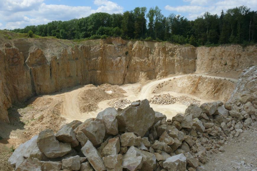 Die unterschiedliche nutzbare Mächtigkeit im Steinbruch Eigeltingen ist auf das Paläorelief zurückzuführen Blick von schräg oben in einen Steinbruch. Das anstehende Gestein ist gelblich-beige. Die Höhe der Abbauwand nimmt von links nach rechts ab, im rechten Teil wächst Wald über der Wand. Im Vordergrund des Bildes liegt Geröll.