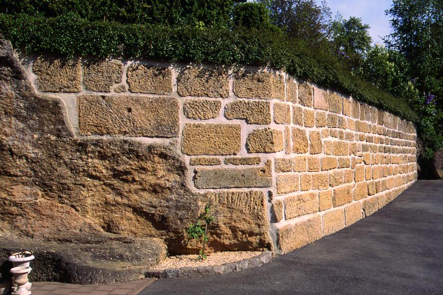 Blick auf die Ecke einer Gartenmauer aus beigen Gesteinsquardern. Im linken Teil besteht die Mauer in der unteren Hälfte aus Felsen, schräg darüber beginnt erst die Mauer. Rechts neben der Mauer befindet sich eine Straße, über der Mauer wächst eine Hecke.