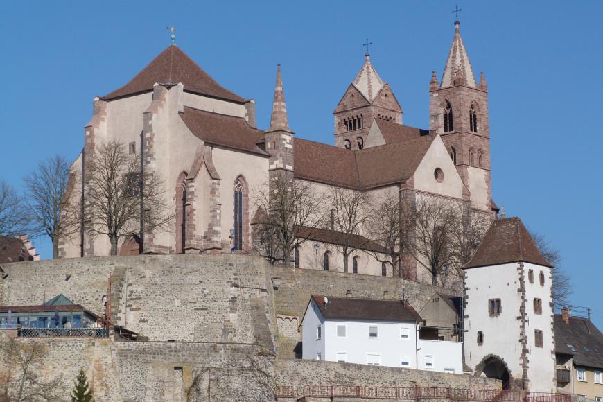 Blick auf eine Kirche mit hellbraunem Mauerwerk und roten Dächern. Links ist ein breiter, viereckiger Turm erkennbar, rechts zwei unterschiedliche, höhere Türme. Die Kirche thront auf einem Mauerberg. Unten rechts steht ein weißer Torturm.