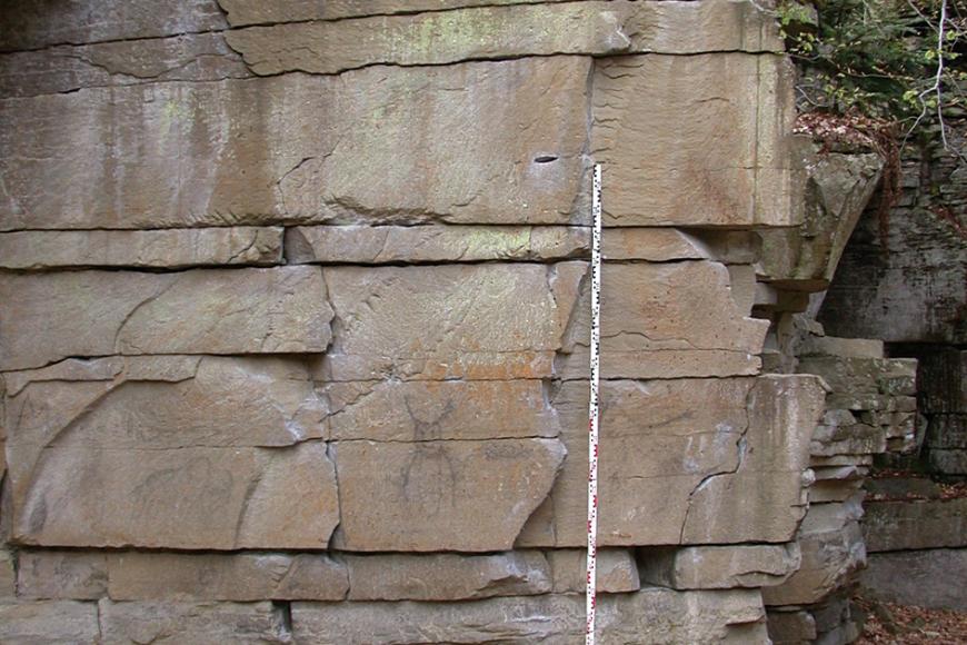 Teilansicht einer hellbraunen Steinbruchwand mit grauem Sockel sowie grünlichen Verfärbungen im oberen Bereich. Der Sockel des Gesteins zeigt zudem Einkerbungen.