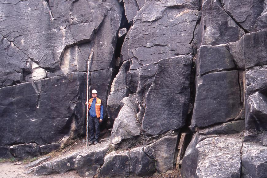 Blick auf eine alte Steinbruchwand mit violettgrauen, massigen Blöcken. Das Gestein ist von zahlreichen Rissen und senkrechten Klüften durchzogen. Links der Bildmitte hält ein Mann in Schutzkleidung eine Messlatte an das Gestein.