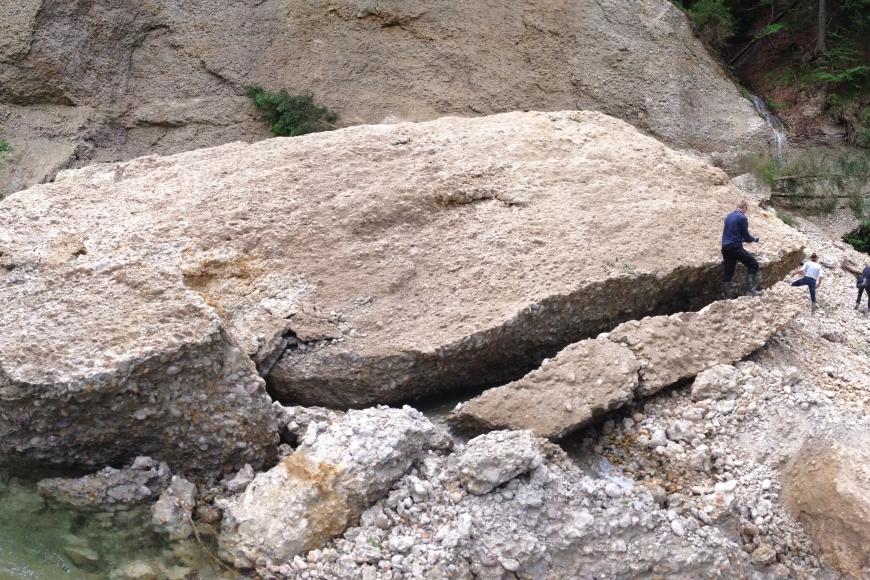 Sturzmasse im Bachbett der Eschach (Foto: WWA Kempten, 2015) Zu sehen ist ein sehr großer und mehrere kleinere Felsblöcke, die vor einer Felswand liegen. Vor den Felsblöcken fließt ein Bach. Rund um den Gesteinsblock herum sind einige Personen zu erkennen, die den mächtigen Block bestaunen.