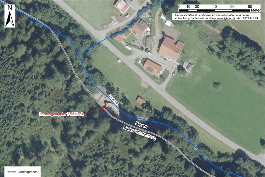 Übersicht des Schadensbereichs mit herausgekipptem Felsblock im Bachbett der Eschach Luftaufnahme eines Baches, in den ein großer Felsblock gekippt ist. Der Bach fließt an einem Waldrand entlang. Rechts und oben sind Wiesen und Häuser erkennbar. In das Luftbild wurden ergänzende Texte und Grenzen eingefügt.