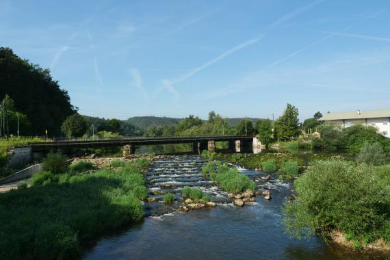 Blick auf einen Flusslauf, der zum Betrachter hin über mehrere flache Stufen hinabfließt. Eine schmale Eisenbahnbrücke überquert den Fluss.