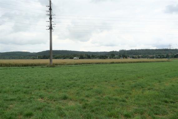 Blick auf flache Wiesen und Äcker. Im Hintergrund, vor langgestreckten bewaldeten Berghängen, verläuft eine stark befahrene Straße.