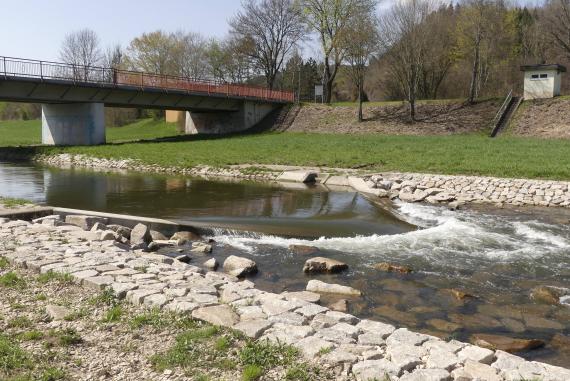 Blick auf einen schmalen Flusslauf mit flacher Staustufe und Strudelbildung im rechten Bildteil. Die Fließrichtung ist von links nach rechts. Der Fluß ist mit Steinufern befestigt. Im Hintergrund links ist eine Brücke, rechts ein Damm mit Bäumen sichtbar.