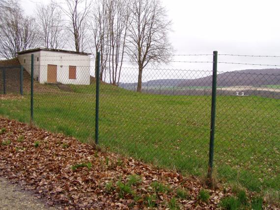 Brunnen Zwiefaltendorf: Fassungsbauwerk im umzäunten Fassungsbereich (Wasserschutzgebietszone 1) Blick auf ein kleines Gebäude mit hellem Verputz und Flachdach. Das Gebäude steht auf einer umzäunten Wiese und ist an einen mit Bäumen bewachsenen Hügel gebaut.