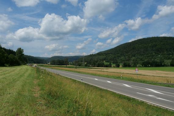 Blick auf flache bis leicht wellige Wiesen und Äcker, die von einer Straße durchschnitten werden. Links und rechts der Straße sowie im Hintergrund erheben sich bewaldete Bergrücken.