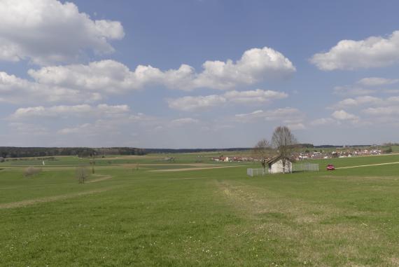 Blick über eine flachwellige Wiesenlandschaft mit Wäldern am Horizont. Im rechten Bildteil, vor einer kleinen Siedlung, steht ein einzelnes eingezäuntes kleines Haus.