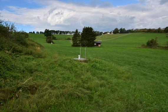 Blick auf welliges Grünland, durchsetzt mit Bäumen. Im Vordergrund, am Beginn eines nach links ansteigenden bewachsenen Hügels, ist ein metallener Schachtdeckel zu sehen.