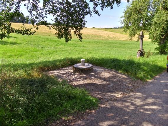 Am Wegesrand einer nach links aufsteigenden, hügeligen Wiese ist auf einer Freifläche ein Schacht eingelassen. Ein Metalldeckel verschließt den Schacht.
