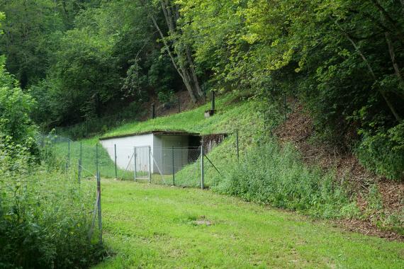Blick auf ein kleines weißes Gebäude ohne Fenster, mit begrüntem Flachdach. Das Gebäude steht vor einem nach rechts aufsteigenden bewaldeten Hang und ist eingezäunt.