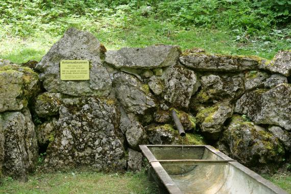 Blick auf eine aus Felsblöcken bestehende Mauer. Aus einem Rohr in der Mauer fließt Wasser in eine ehemalige Viehtränke. Links der Bildmitte ist eine Schrifttafel erkennbar; auf ihr steht „Antoniusbrunnen“..