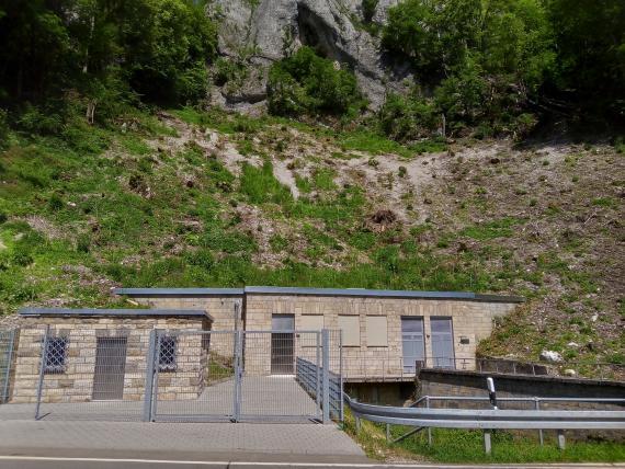 Blick auf mehrere eingezäunte Gebäude mit Flachdächern und teils vergitterten Fenstern. Die Gebäude stehen vor einem teilweise gerodeten Steilhang, an dessen oberem Ende Felsgestein zu erkennen ist.