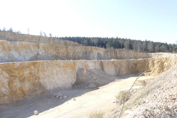 Liegende Bankkalke-Formation im Schotterwerk Baur, Veringenstadt-Jungnau Seitlicher Blick in einen Steinbruch. Zu sehen sind links drei lange Gesteinsstufen sowie rechts ein Abraumhaufen. Dazwischen ist der flache Boden des Steinbruchs sichtbar.