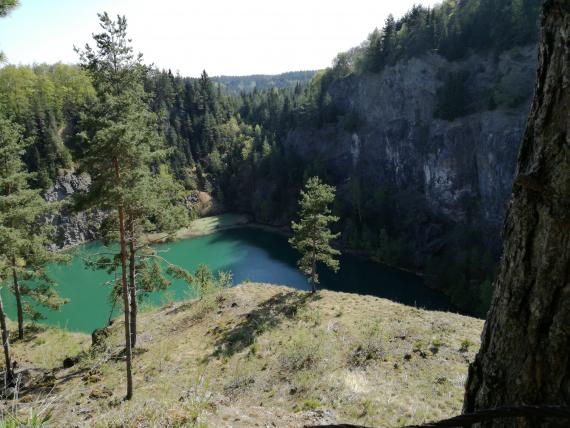 Nach unten, über ein Felsplateau gerichteter Blick auf einen grünen See. Das Ufer links des Sees besteht aus Felsen und Wald, das Ufer rechts aus einer hohen, im Schatten liegenden Felswand.