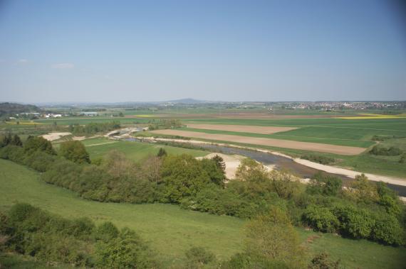 Blick von der Keltensiedlung Heuneburg über das Donautal zum Bussen Blick über Hangstufen mit Grünstreifen und Bäumen auf eine weite Landschaft mit Äckern und Wiesen. Ein Fluss mit flachen, hellen Ufern durchzieht die Landschaft. Im fernen Hintergrund ist rechts eine Siedlung und über der Bildmitte ein Bergkegel sichtbar.
