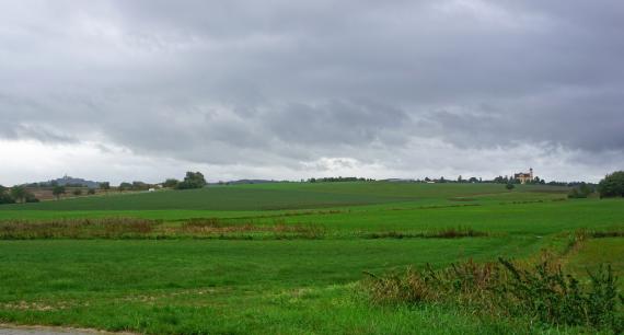 Das Bild zeigt in einer Senke liegende Feuchtwiesen. Zum Mittel- und Hintergrund hin folgen flach ansteigende Hügel mit weiterem Grünland. Rechts auf einer Hügelkuppe steht eine Kirche. Links hinten erhebt sich ein bewaldeter Schlossberg.