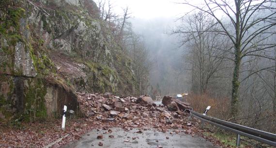 Im Vordergrund befindet sich eine Straße, die von Blöcken und Steinen bedeckt ist. Links befindet sich eine steile Flanke, an der Festgestein ansteht.