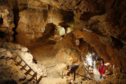 Blick von erhöhtem Standpunkt in eine große Höhlenkammer. Die Wände hinten und rechts bestehen aus bräunlichem, teils beleuchtetem Gestein. Links führt eine Steintreppe aufwärts, rechts ein schmaler Gang abwärts.