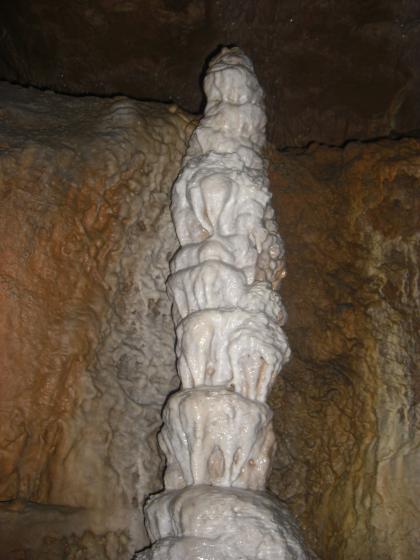 Blick in eine Höhle mit einem säulenartigen, von unten nach oben gewachsenen Tropfstein. Weißlich gefärbt, hebt er sich gut von den dunkelbraunen Höhlenwänden dahinter ab.