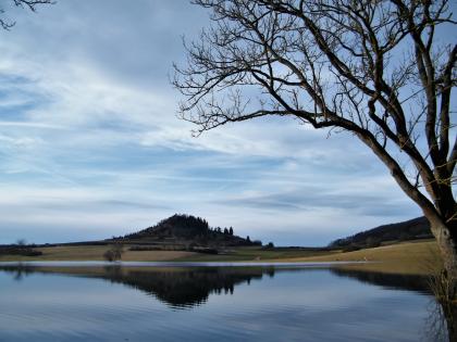 Blick auf einen ruhigen See. Im dahinter liegenden Uferbereich erhebt sich links der Bildmitte ein bewaldeter, kegelförmiger Berg. Rechts ragt ein kahler Baum ins Bild.