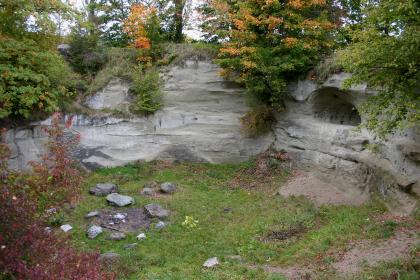 Blick von oben auf eine halbrunde, graue Gesteinswand, in der rechts eine Vertiefung ausgehöhlt wurde. Am Boden vor der Gesteinswand ist eine Feuerstelle. Die oberen Ränder des Gesteins sind bewachsen.