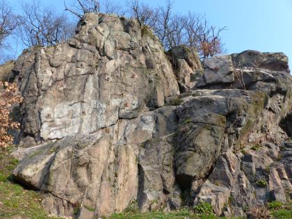 Blick auf ein klippenartige Felsgruppe aus graugrünen Gesteinsblöcken. Die erhöhte Kuppe links ist von Bäumen bewachsen.