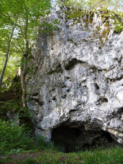Blick auf einen grauen, turmartigen Felsblock, der zum linken Bildrand hin steil abfällt. Am Fuß des Felsens, der oben bemoost und bewachsen ist, öffnet sich in flachem Bogen eine Höhle.