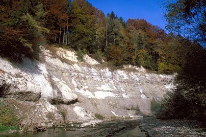 Am linksseitigen Ufer eines schmalen Flusses erhebt sich eine hohe, lange Felsböschung. Der Fels ist grau mit weißen Flecken und teils glatt, teils hervorstehend. Die Kuppe der Böschung ist bewaldet.