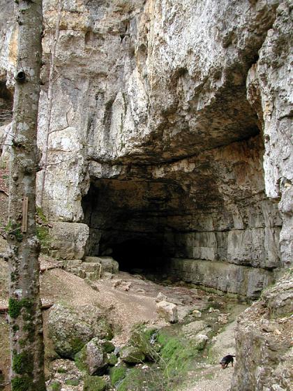 Blick auf den Eingang zu einer Höhle, am unteren Ende einer grauen, fast senkrecht aufragenden Felswand. Ein schmaler Pfad führt rechts an einem Felsenhang vorbei zur Höhle. 
