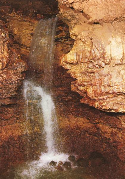 Man sieht das Innere einer Höhle mit stufigem Wasserfall links und diesen eng umschließende, rötlich braune Gesteinswände.