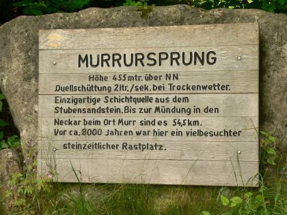 Informationstafel aus Holz am Murr-Ursprung, befestigt an einer größeren Steinplatte. Die Tafel gibt Auskunft etwa über das Schüttungsvolumen oder die Länge der Murr bis zu ihrer Vereinigung mit dem Neckar.
